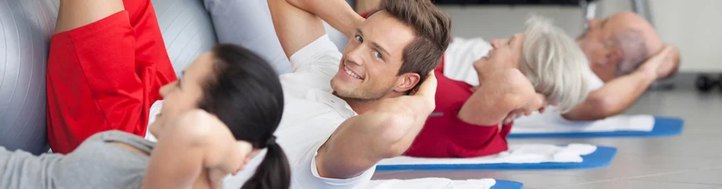 Vier Personen unterschiedlichen Alters liegen auf Übungsmatten und machen Bauchmuskelübungen in einem Fitnessstudio oder einem Fitnesskurs. Ein Mann in der Mitte lächelt in die Kamera. Bild im 123fit Rahlstedt