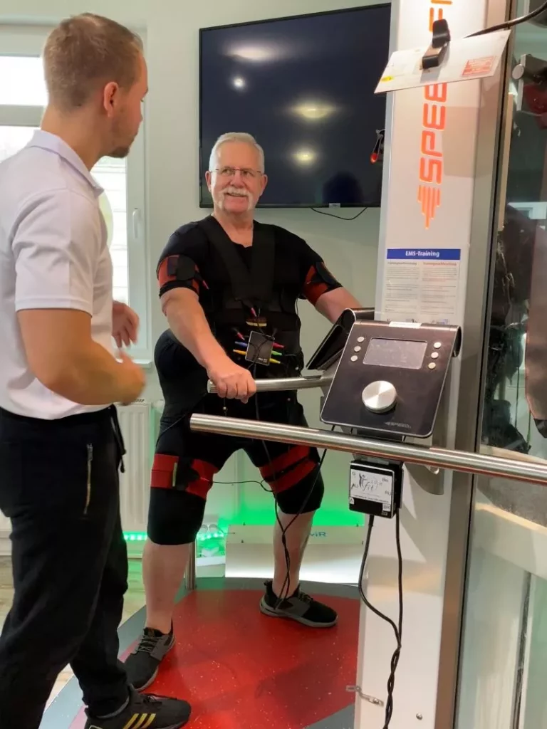 Ein älterer Mann in einem EMS-Trainingsanzug hält sich an den Griffen eines Fitnessgeräts fest, während ein Trainer in einem weißen Hemd ihn in der Halle anleitet. Ein Monitor und elektronische Geräte sind zu sehen. Bild im 123fit Rahlstedt