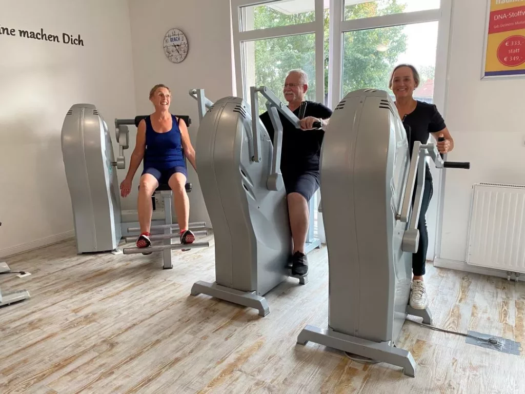 Drei Erwachsene trainieren an sitzenden Trainingsgeräten in einem hellen Fitnessraum mit Holzboden und großen Fenstern. Die Personen wirken konzentriert und sind sportlich gekleidet. Bild im 123fit Rahlstedt
