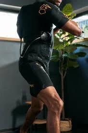 Eine Person, die einen schwarzen Hightech-Anzug mit gelben Akzenten trägt, steht in einem Innenraum und scheint eine körperliche Tätigkeit auszuüben. Im Hintergrund sind eine große Pflanze und ein Fenster zu sehen. Bild im 123fit Rahlstedt