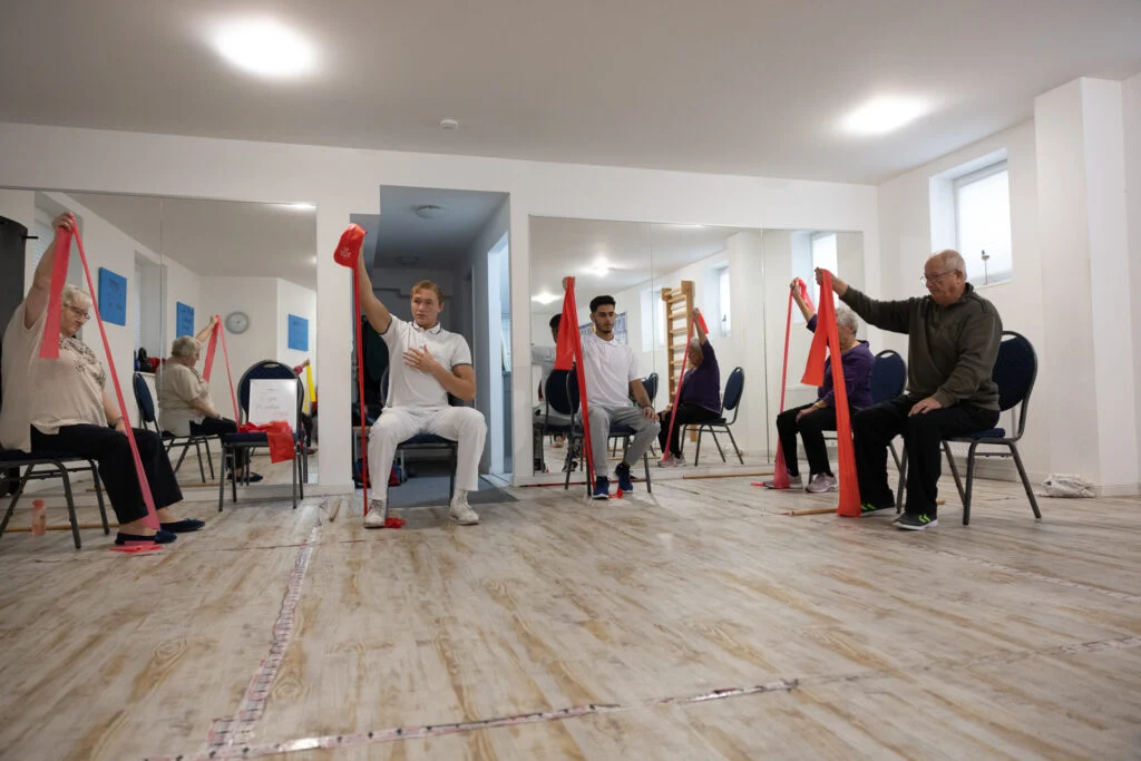 Vier Personen sitzen auf Stühlen in einem Raum mit Spiegeln und halten jeweils mit einer Hand ein rotes Übungsband über dem Kopf. Sie scheinen an einem Gruppenfitness- oder Rehabilitationskurs teilzunehmen. Bild im 123fit Rahlstedt