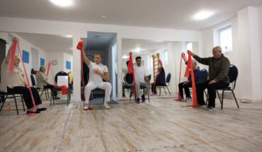 Vier Personen sitzen auf Stühlen in einem Raum mit Spiegeln und halten jeweils mit einer Hand ein rotes Übungsband über dem Kopf. Sie scheinen an einem Gruppenfitness- oder Rehabilitationskurs teilzunehmen. Bild im 123fit Rahlstedt