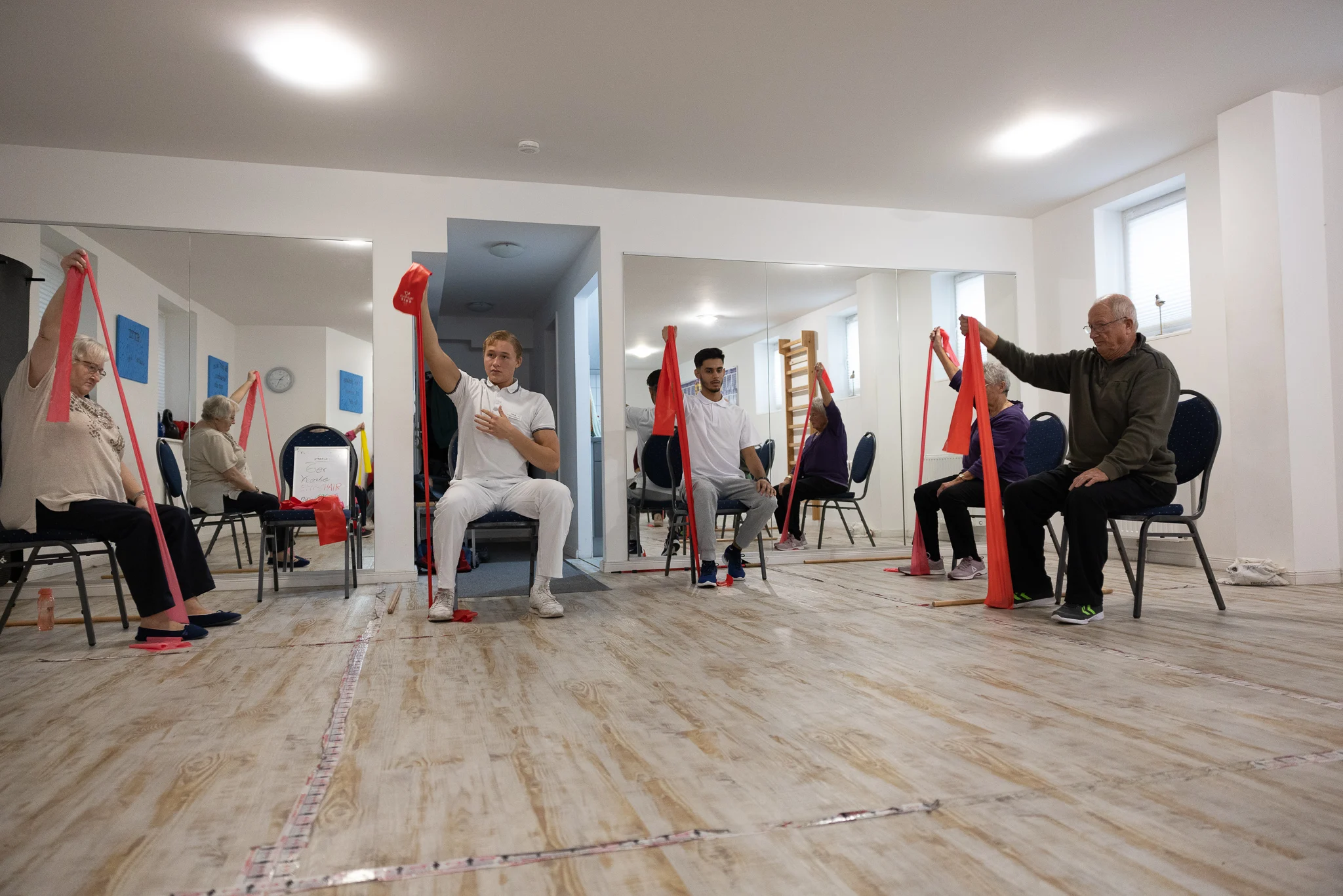 Vier Personen sitzen auf Stühlen in einem Raum mit Spiegeln und halten jeweils mit einer Hand ein rotes Übungsband über dem Kopf. Sie scheinen an einem Gruppenfitness- oder Rehabilitationskurs teilzunehmen. Bild im 123fit Rahlstedt