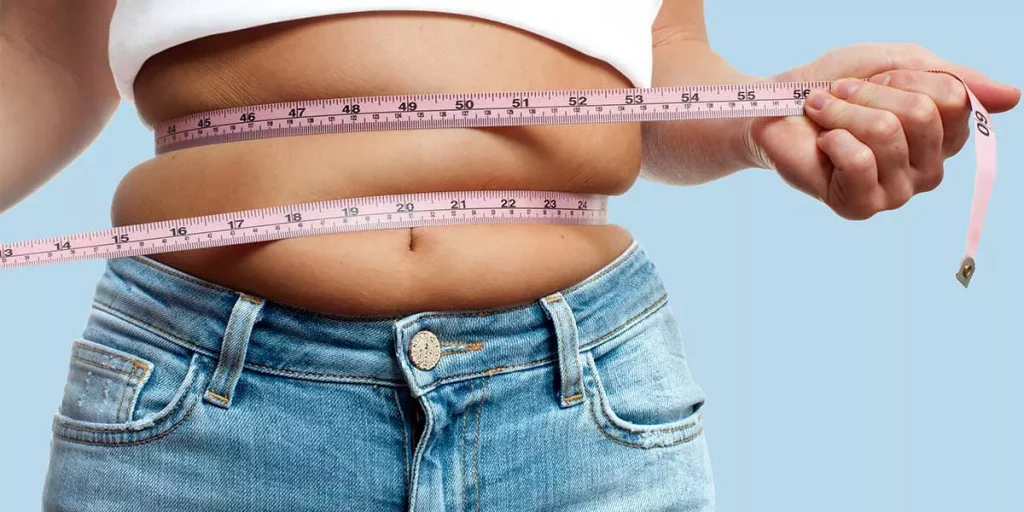 Eine Person, die Jeans und ein weißes Oberteil trägt, misst ihre Taille mit einem rosa Maßband. Das Band ist um die Körpermitte gewickelt, und die Hand hält es fest. Der Hintergrund ist hellblau. Bild im 123fit Rahlstedt