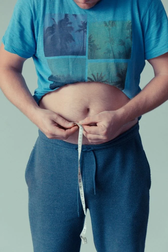 Eine Person, die ein blaues T-Shirt und eine Jogginghose trägt, misst ihre Taille mit einem Maßband und entblößt dabei ihren Bauch. Das Gesicht der Person ist nicht zu sehen. Bild im 123fit Rahlstedt